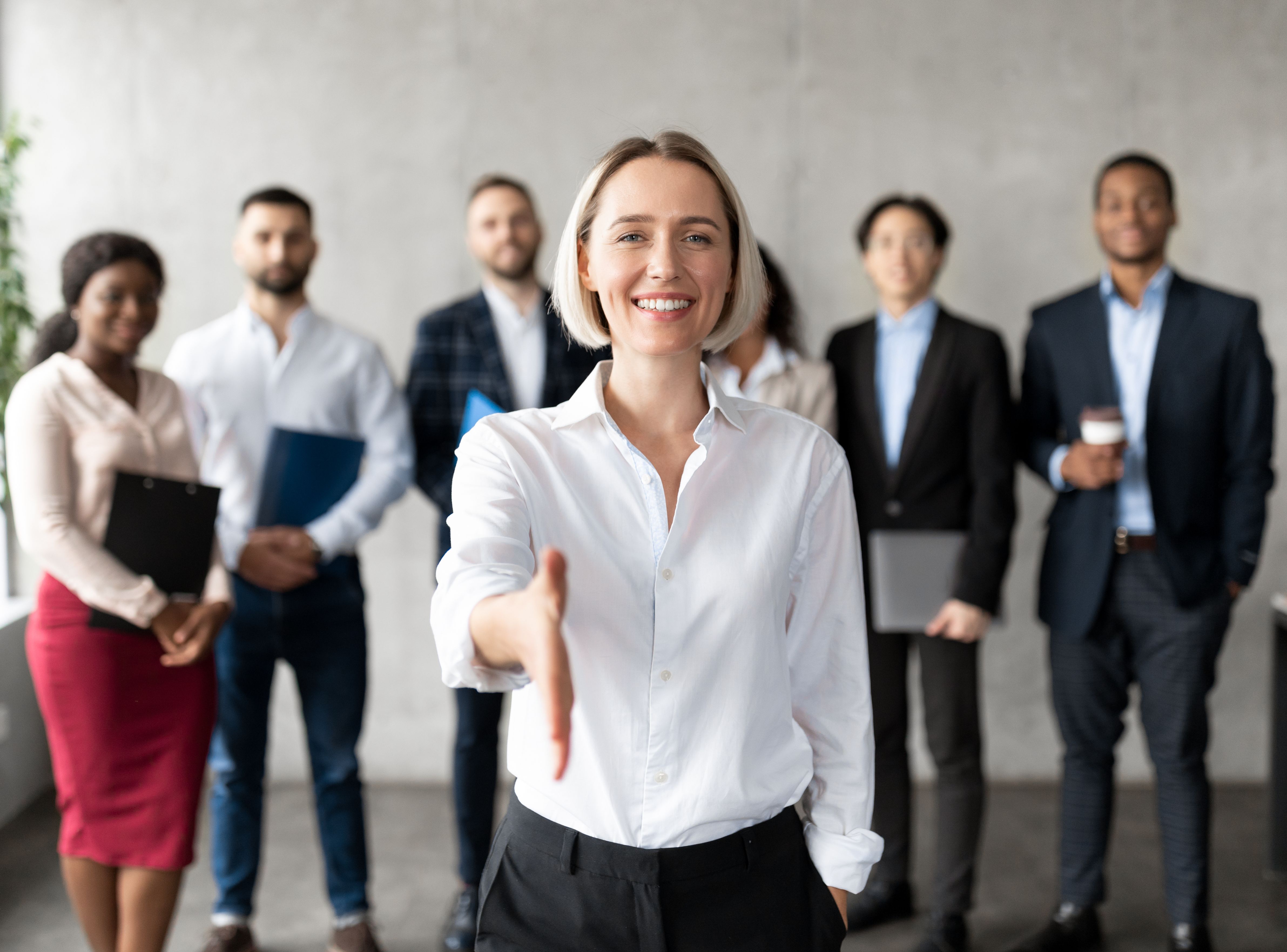 Blonde Frau im Business-Outfit reicht zur Begrüßung die Hand, im Hintergrund steht das Team der Zeitarbeitsfirma und heißt zum Zeitarbeit-Job Stuttgart willkommen.